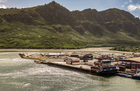 Nawiliwili, Kauai, Hawaii, USA. - January 17, 2020: Almost empty Haaheo shipping container barge docked in port. Green mountain with brown-black cliffs as backdrop under light blue cloudscape.のeditorial素材