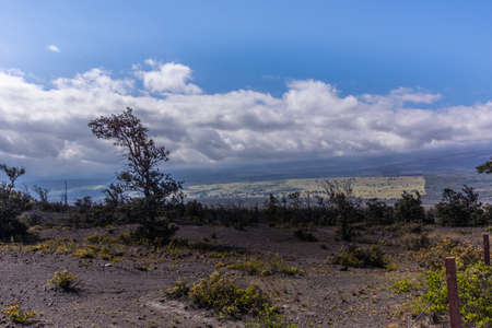 Kilauea volcano, Hawaii, USA. - January 9, 2012: Looking down from the mountain onto surrounding land under blue cloudscape. Black lava rock with some green vegetation up front.の写真素材