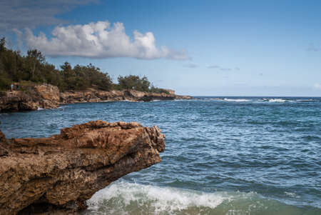 Kauai, Hawaii, USA. - January 11, 2012: Brown rough rocky coastline at Kawailoa bay with blue ocean, blue sky and white clouds. Green trees on top of cliffs. Hidden fisherman.の写真素材