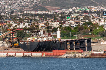 Ensenada, Mexico - January 17, 2012: Black barge Toltec in dry dock in port. Backed by white cityscape on slope of mountain. Blue bay water in front.のeditorial素材