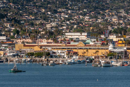 Ensenada, Mexico - January 17, 2012: Coca-Cola billboard above yellow builidngs on quay if the Marina with small yachts and other vessels on blue bay water and cityscape behind on slope of hill.のeditorial素材