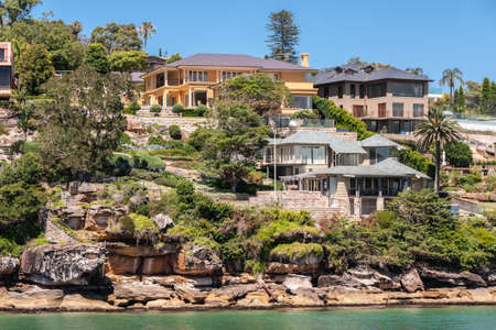 Sydney, Australia - December 11, 2009: Upscale housing with gray roofs on rocky slope with green vegetation on northern shore  of bay under blue sky and behind green bay water.のeditorial素材