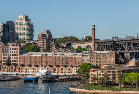 Sydney, Australia - December 11, 2009: Historic brown brick warehouses of Rocks neighborhood with on-ramp of Harbour Bridge in back under blue sky. Boat and some green foliage.のeditorial素材