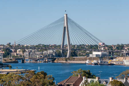 Sydney, Australia - December 11, 2009: 1 of 2 suspension pylons of ANZAC Bridge set in cityscape under light blue sky and behind deep blue water. Some green foliage.のeditorial素材