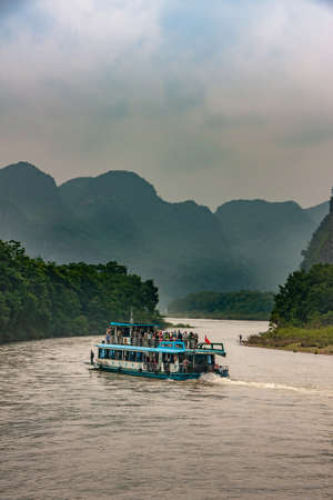 Guilin, China - May 10, 2010: Along Li River. White-blue overloaded tourist sightseeing boat meanders with brown water Green shoreline and karst mountains under brown foggy sky.のeditorial素材