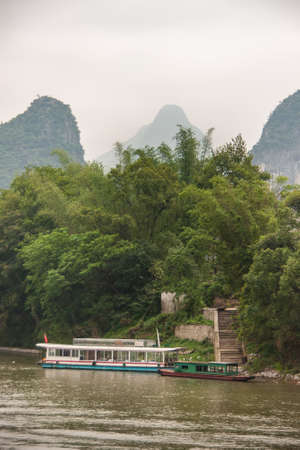 Guilin, China - May 10, 2010: Along Li River. Ferry and transport vessel on brown water docked at stairs surrounded by green foliage with karst mountain tops in foggy gray sky as backdrop.のeditorial素材