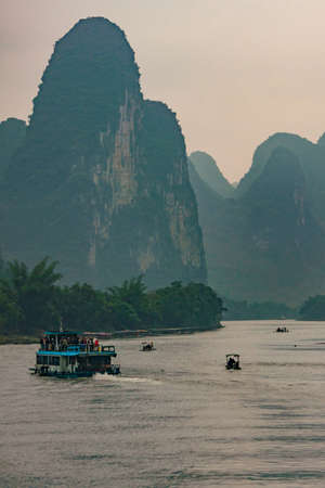 Guilin, China - May 10, 2010: Along Li River. Overloaded blue sightseeing ferry and three very small vessels sailing in gorge of tall karst mountains with cliffs under foggy sky.のeditorial素材