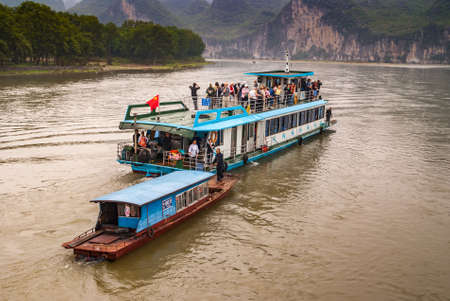 Guilin, China - May 10, 2010: Along Li River. Vendor on small boat approaches on brown water blue-white tourist sightseeing vessel. Rocky cliffs and green foliage as backdrop.のeditorial素材
