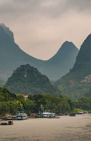 Guilin, China - May 10, 2010: Along Li River. Busy stretch with multiple boats along green forested shoreline. Tall karst mountains covered in green under gray cloudscape as backdrop.のeditorial素材