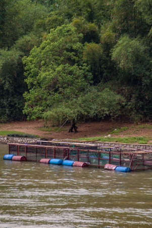Guilin, China - May 10, 2010: Along Li River. Closeup of Blue and red metal, barrel-supported fish pen near shoreline full of green trees.のeditorial素材
