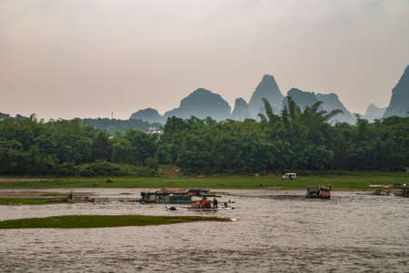 Guilin, China - May 10, 2010: Along Li River. On wide stretch of river with banks, house boat, rafts and ferries in front of green shoreline with tall karst mountains under silver sky as backdrop.のeditorial素材