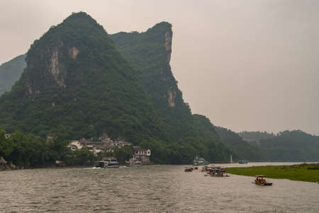 Guilin, China - May 10, 2010: Along Li River. Boat, raft and ferry traffic around the bend with green karst mountain top and vertical cliffs, houses at foot, as backdrop.のeditorial素材