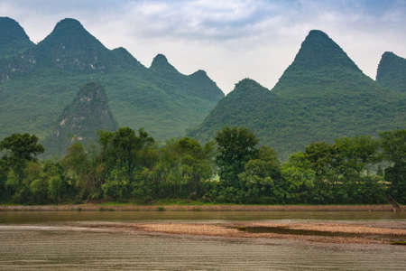 Guilin, China - May 10, 2010: Along Li River. Landscape with chain of green forested karst mountain and under blue cloudscape. Green tree belt, Brown water and marsh land up front.の写真素材
