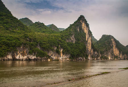 Guilin, China - May 10, 2010: Along Li River. Landscape of karst mountain under blue cloudscape with brown-black vertical cliff. Land tongue extension dirt line in dark brown water.の写真素材