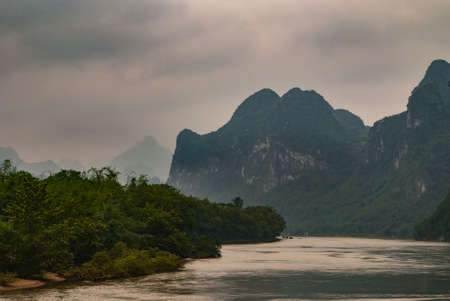 Guilin, China - May 10, 2010: Along Li River. Landscape of dark green covered karst mountains under gray cloudscape. Small boats on horizon on silver river. Green foliage on shoreline.の写真素材