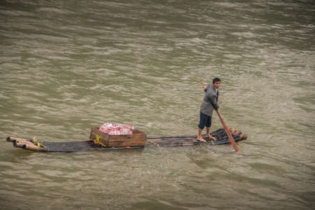 Guilin, China - May 10, 2010: On Li River. Closeup, Male vendor with gray vest on his flimsy raft on green choppy water. Brown crate with red-white bag of merchandise.のeditorial素材
