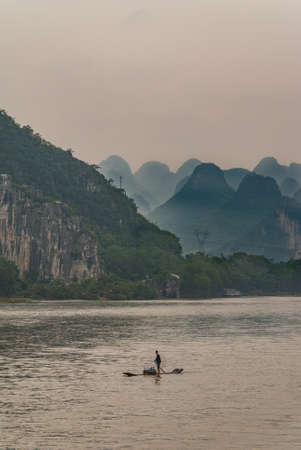 Guilin, China - May 10, 2010: On Li River. Wide shot with Male vendor with on his flimsy raft on silver water. Brown crate with merchandise. Karst mountains and forest as backdrop. Gray sky.のeditorial素材