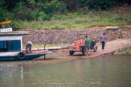 Guilin, China - May 10, 2010: Along Li River. People manually load red bricks from old pickup truck into barge on a slipway. Green forest in back. Greenish water up front.のeditorial素材