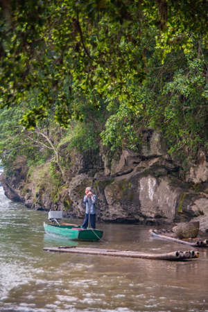 Guilin, China - May 10, 2010: Along Li River. Lone man in blue garb stands in green sloop near dark gray rocky shoreline and under green foliage. Some flimsy rafts nearby on brown greenish water.のeditorial素材