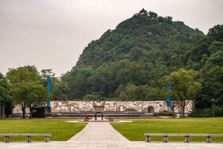 Guilin, China - May 11, 2010: Seven Star Park. Modernist Nationalist memorial wall in front of green covered mountain under silver sky. In front: stage, offering vessel, mother and child and green lawn.のeditorial素材