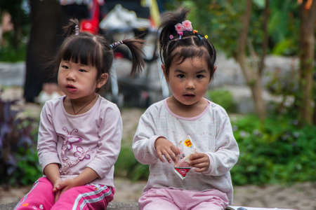 Guilin, China - May 11, 2010: Seven Star Park. Frontal closeup of 2 little girls sitting on bench. Fake flowers in black hair. Light shirts and multicolor backdrop.のeditorial素材