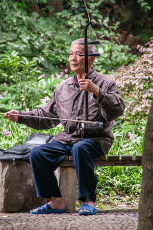 Guilin, China - May 11, 2010: Seven Star Park. Man sitting on bench plays the erhu or Chinese violin. Green foliage in back.のeditorial素材