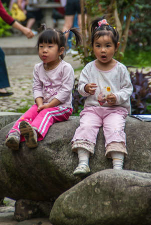 Guilin, China - May 11, 2010: Seven Star Park. Frontal closeup of 2 little girls sitting on dark gray boulder. Fake flowers in black hair. Light shirts and multicolor backdrop.のeditorial素材