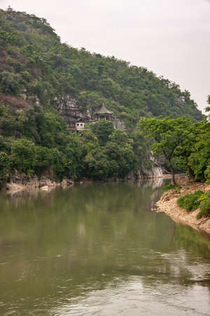 Guilin, China - May 11, 2010: Xiadong River. Chinese traditional brown wood buildings on slope of green covered hill along greenish water.の写真素材