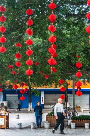 Guilin, China - May 11, 2010: Multiple srings of red balloons hang from tree branches with green foliage. Street scene with people and closed store.のeditorial素材