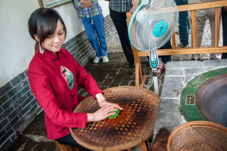 Guilin, China - May 11, 2010: Tea Institute. Young woman in red uniform rolls green leaves on reed plate as preparation for roasting them in a minute on oven in front of her.のeditorial素材
