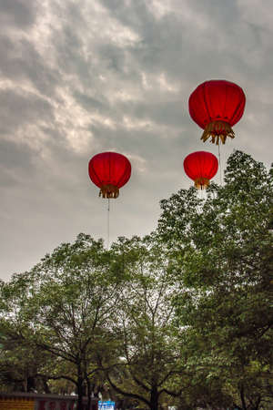 Guilin, China - May 10, 2010: 3 small red hot air balloons with candle inside float above green trees under dark cloudscape.の写真素材