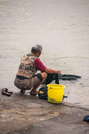 Guilin, China - May 11, 2010: Downtown. Woman in brown and maroon garb does her laundry sqatting at water edge of silver Li River with yellow bucket on her side.のeditorial素材