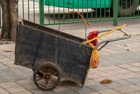 Chongqing, China - May 9, 2010: Downtown. Closeup of Black box push cart to collect trash and garbage in street. Yellow broom stick and red safety vest. Green fence in back.のeditorial素材