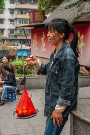 Chongqing, China - May 9, 2010: Downtown. Closeup of young woman in blue garb selling red bag with a few fruits on street corner while holding a simple gray scale. Other people.のeditorial素材