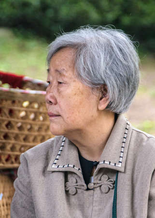 Chongqing, China - May 9, 2010: Inside zoo or animal park. Closeup of serious face of graying woman wearing beige-brown vest against faded green foliage and yellow basket backdrop.のeditorial素材