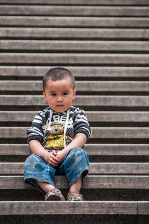 Shanghai, China - May 4, 2010: Closeup of little boy wearing black-white shirt and wide jeans, sitting on brown concrete steps, and looking seriously and observing into the camera.のeditorial素材
