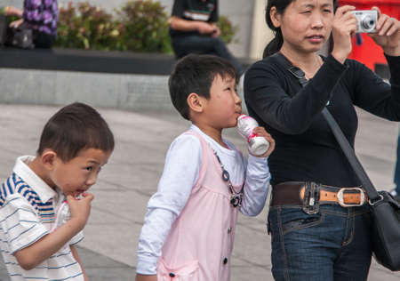 Shanghai, China - May 4, 2010: 2 young boys, wearing light colored clothes, sip yoghurt out of plastic bottle while one also looks as black clad mother takes picture.のeditorial素材