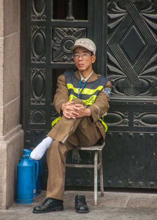 Shanghai, China - May 4, 2010: Front door guard in brown uniform and yellow security vest sits on chair and has one of his black shoes taken off exposing his white sock. Blue water cans next to him.のeditorial素材