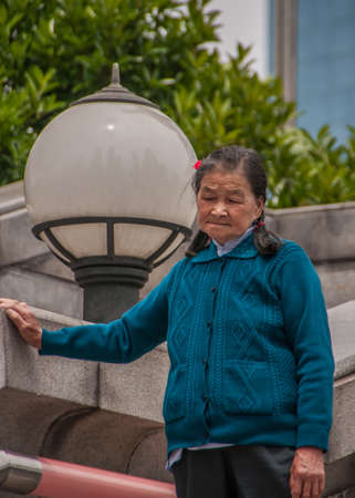 Shanghai, China - May 4, 2010: Older woman in blue vest holds herself steady on gray stone wall. Big ball lantern and green foliage in back.のeditorial素材