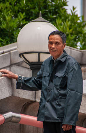 Shanghai, China - May 4, 2010: Old man with bad teeth and in dark blue vest holds himself steady on gray stone wall. Big ball lantern and green foliage in back.のeditorial素材