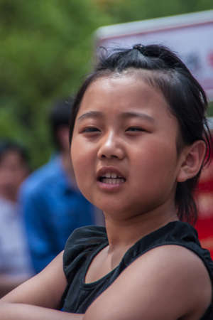 Shanghai, China - May 4, 2010: Closeup of face of defiant young woman, arms crossed, looks straight into the camera. Green foliage in back.のeditorial素材