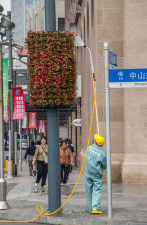 Shanghai, China - May 4, 2010: Male worker in green uniform waters hanging flower pieces along Bund, in front of Citi Bank building. Street scene with pedestrians and banners adding color.のeditorial素材