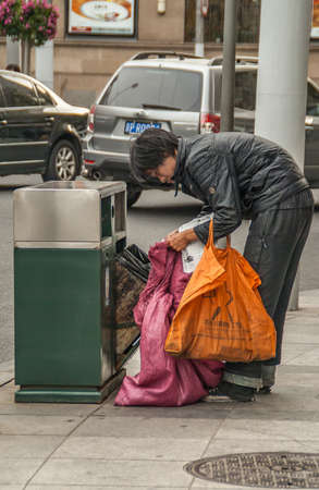 Shanghai, China - May 4, 2010: Relatively young person dressed in black searches public trash can  while holding pink and orange bags. Street scene with cars.のeditorial素材