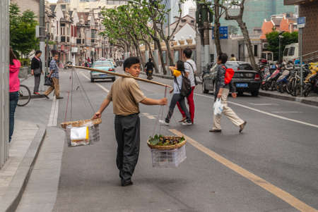 Shanghai, China - May 4, 2010: Man carries baskets filled with mangosteen fruit on wooden shoulder pole over street with other people around and cars .のeditorial素材