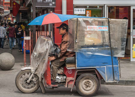 Shanghai, China - May 4, 2010: Closeup of man riding a motor tricycle with umbrella to protect driver and loading compartment with clear plastic cover. Street scene with pedestrians.のeditorial素材
