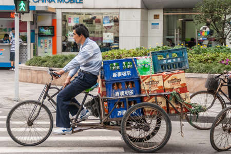 Shanghai, China - May 4, 2010: Closeup of man in shirt and pants riding a pedal tricycle with blue bottle cratesa and brown cartons in loading compartment. Street scene.のeditorial素材