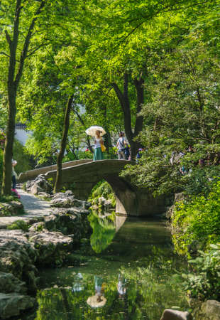 Suzhou China - May 3, 2010: Humble Administrators Garden. Woman with white umbrella stands on gray bridge, both reflected in canal against wall of green foliage. Path along water.のeditorial素材