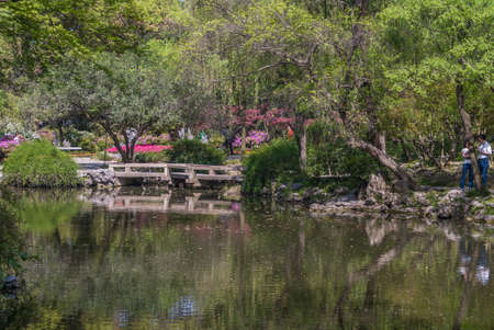 Suzhou China - May 3, 2010: Humble Administrators Garden. Wall of green foliage across greenish mirroring water of canal with small bridge. Red flowers and people sprinkled.のeditorial素材