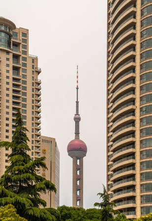 Shanghai, China - May 4, 2010: Oriental Pearl TV tower seen from between yellow-brown skyscrapers against silver sky with some green foliage up front.のeditorial素材