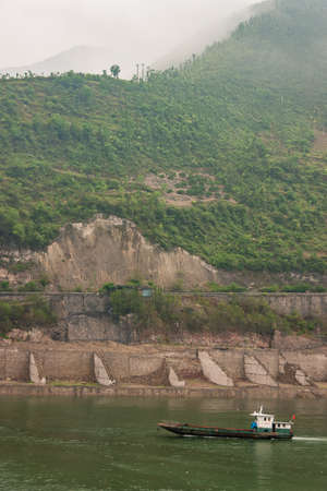 Baidicheng, China - May 7, 2010: Qutang Gorge on Yangtze River. Small boat on green water with green tree covered mountain slope, top in the fog, and cliffs with road near river.の写真素材